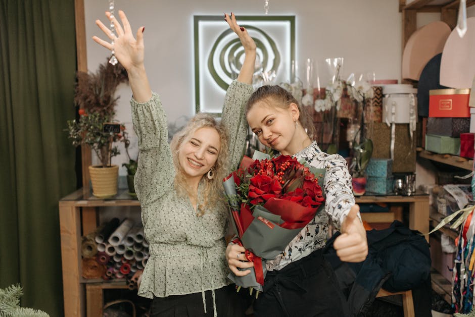 Two young women standing in a florist shop, one holding a large bouquet of red roses wrapped in grey paper with a red ribbon, both smiling and appearing happy. The woman on the left has curly blonde h
