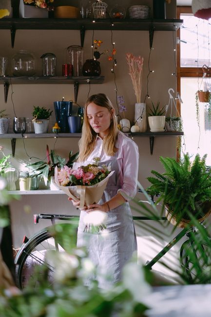 A young woman with long, light brown hair dressed in a white shirt and apron is holding a fresh floral bouquet wrapped in beige paper. She is standing inside a bright, well-lit flower shop with shelve