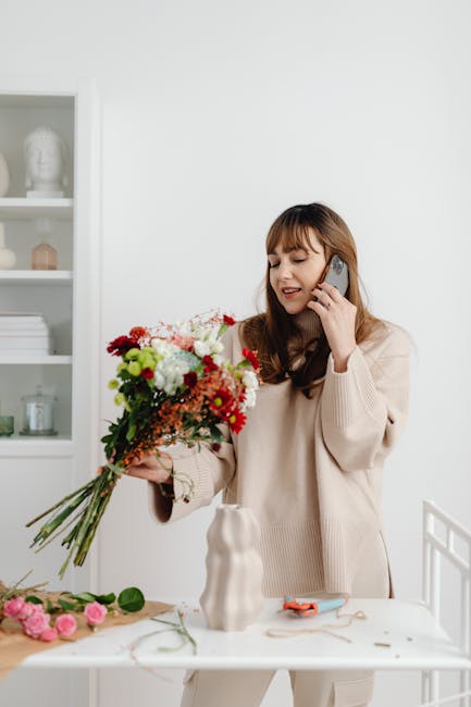 A young woman with shoulder-length brown hair and light skin, wearing a beige sweater, is smiling slightly while talking on a mobile phone held to her ear. She is holding a large, vibrant bouquet of f