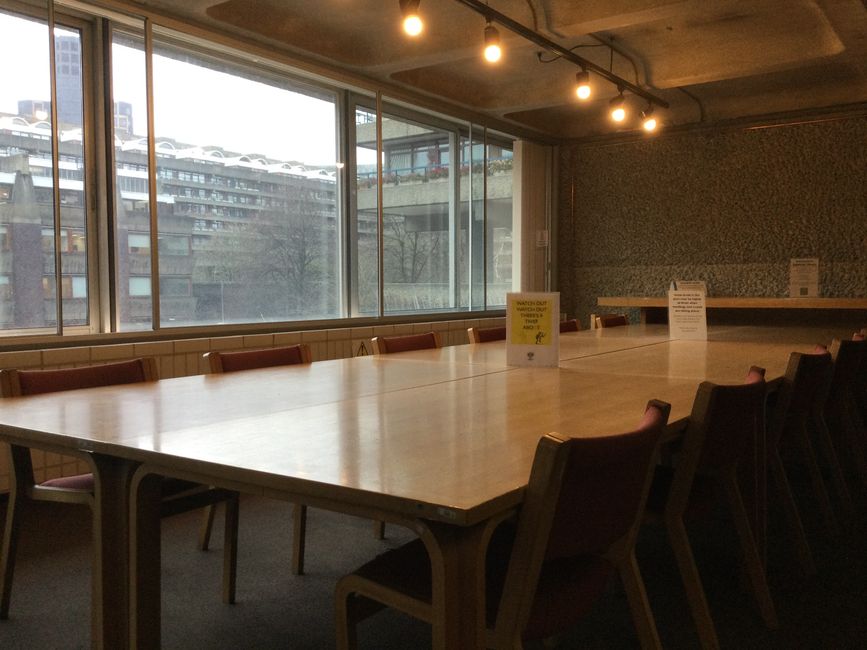 Inside a modern conference room with a long, rectangular wooden table surrounded by multiple chairs with upholstered seats. The room features large floor-to-ceiling windows that allow natural light to