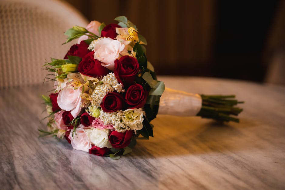 A close-up of a romantic bouquet arrangement featuring deep red roses, soft pink ranunculuses, white lisianthus, and delicate sprigs of white gypsophila, all arranged with green foliage. The flowers a