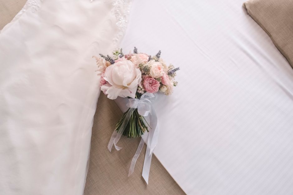 A delicate bouquet of fresh flowers arranged with soft pink and white peonies, roses, and lavender sprigs, wrapped in elegant white tissue paper and tied with a satin ribbon. The bouquet is resting on