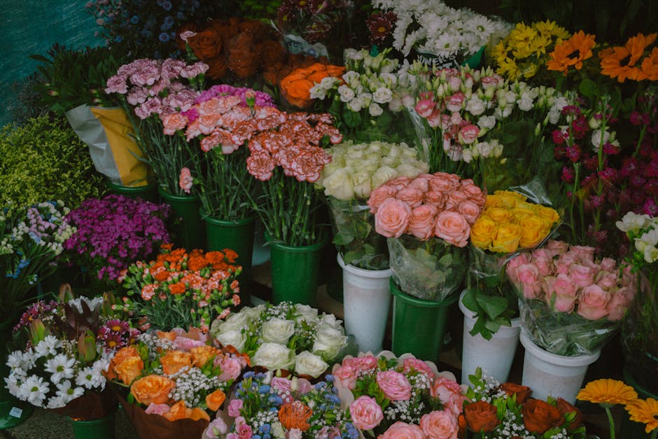 An assortment of fresh floral arrangements and bouquets displayed in green and white vases and wrapping, featuring a variety of flowers including pink and peach roses, white roses, orange and yellow b