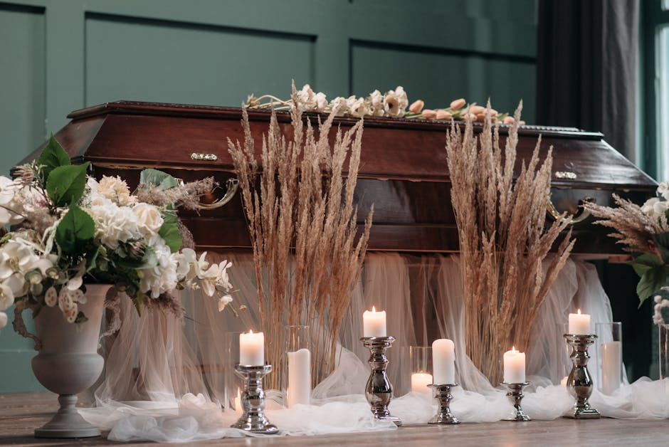 An arrangement of fresh white and blush pink flowers, including roses and hydrangeas, displayed in elegant white vases on either side of a wooden casket. Tall, beige dried pampas grass stalks are posi