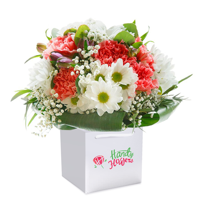 Set against a luminous white background, a generous floral bouquet blooms from within a modern, square white gift bag-its style and freshness echoing the contemporary vibrancy of White City. Soft coral-pink and deep red carnations take center stage, their plush, ruffled petals layered in a display of texture and warmth. Dazzling white daisies, each with an expressive green-gold center, punctuate the arrangement, providing a crisp contrast and a sense of lightness. Airy clouds of white gypsophila sprinkle throughout, enhancing the bouquet's delicate structure. Peeking on the left, unopened buds subtly veined with purple introduce depth and anticipation, reminiscent of the wildflowers spotted along the edges of Hammersmith Park. At the bouquet's base and framing the arrangement, sleek glossy leaves arc outward, creating a rich, verdant collar-suggestive of the flourishing greenery woven through White City's streets. The ensemble forms a beautifully rounded shape, projecting both freshness and generosity of spirit. Completed by the gift bag's ribbon handles for handy carrying, this bouquet stands ready to celebrate everything from birthdays to reunions in the heart of White City, its every detail reflecting the artistic eye and warm heart of the local florist community.