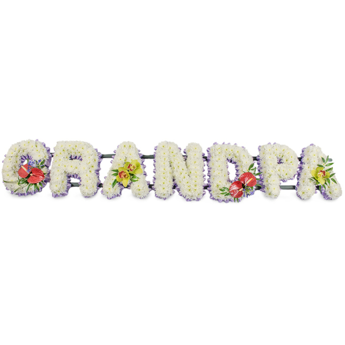 The photograph shows a refined and emotionally resonant funeral tribute spelling out "GRANDPA" in substantial, upright letters that run horizontally from one side of the image to the other. Every letter is built upon a sturdy frame but is completely blanketed in small, fresh white chrysanthemums, giving the word a soft, continuous texture, almost like a row of floral cushions. The chrysanthemums are placed closely together so no greenery shows through, their layered petals catching gentle highlights and shadows that add depth to the arrangement. A slim trim of pale lavender ribbon runs along the base of each letter, the muted purple hue echoing tones that often feel at home in quiet chapels and crematoriums around White City and the wider Hammersmith and Fulham area. This ribbon border neatly defines the bottom edge of each letter without disturbing the clean, white purity of the mums. Behind the flowers, dark green bars provide the structural link between letters, holding the tribute steady whether it rests atop a coffin or stands on easels for a memorial service. Four carefully composed accent sprays punctuate the design. On the 'G' and 'D', clusters of rich red anthuriums emerge from the chrysanthemums, their waxy, heart-shaped surfaces and central spadices framed by glossy green leaves and the occasional tiny filler flower, giving a sense of intensity and heartfelt remembrance. By contrast, the first and final 'A' display graceful yellow-green orchids, their curved petals, intricate markings, and subtle lime tones suggesting gentle admiration and enduring affection. Together, the interplay of white, red, lavender, and green creates a balanced, dignified composition. It feels like the work of a local florist in White City, thoughtfully crafted for a family wishing to honour their grandfather with a personal, meaningful floral message of love and farewell.